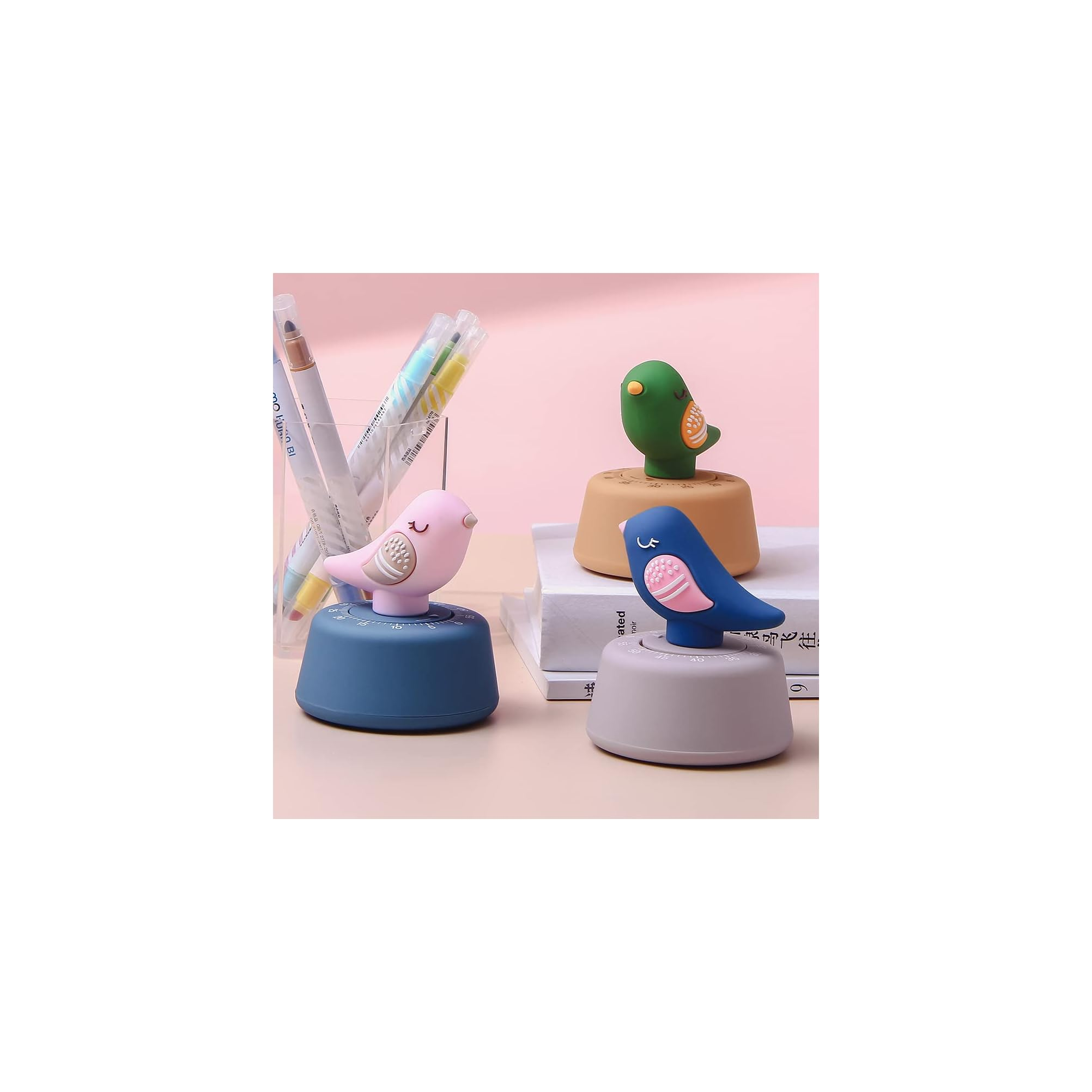 Drei bunte, vogelörmige Küchenwecker auf einer rosa Oberfläche mit Stiftehalter und Büchern im Hintergrund.