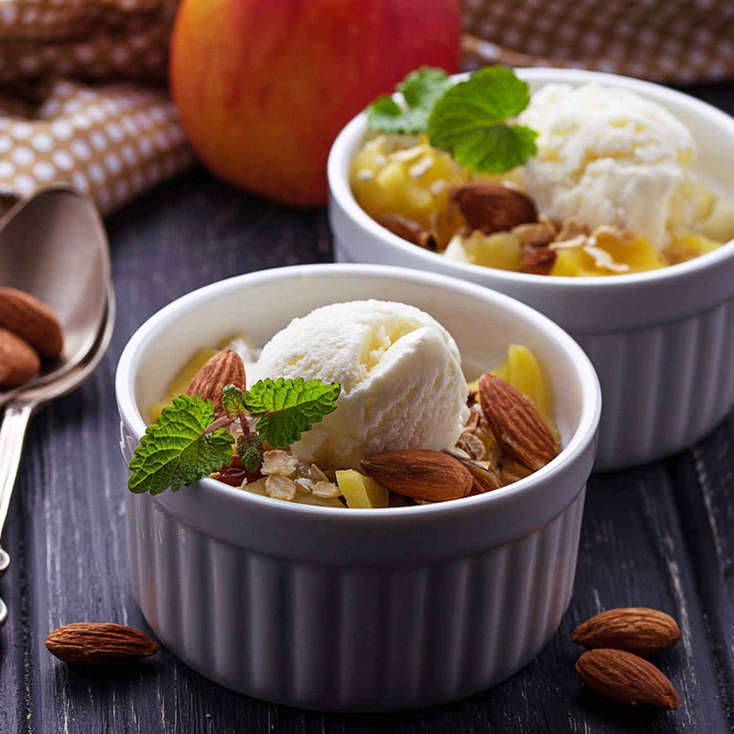 Zwei weiße Schalen mit Eis, Mandeln, Obst und Minze auf dunklem Holz, mit Apfel und Löffel.