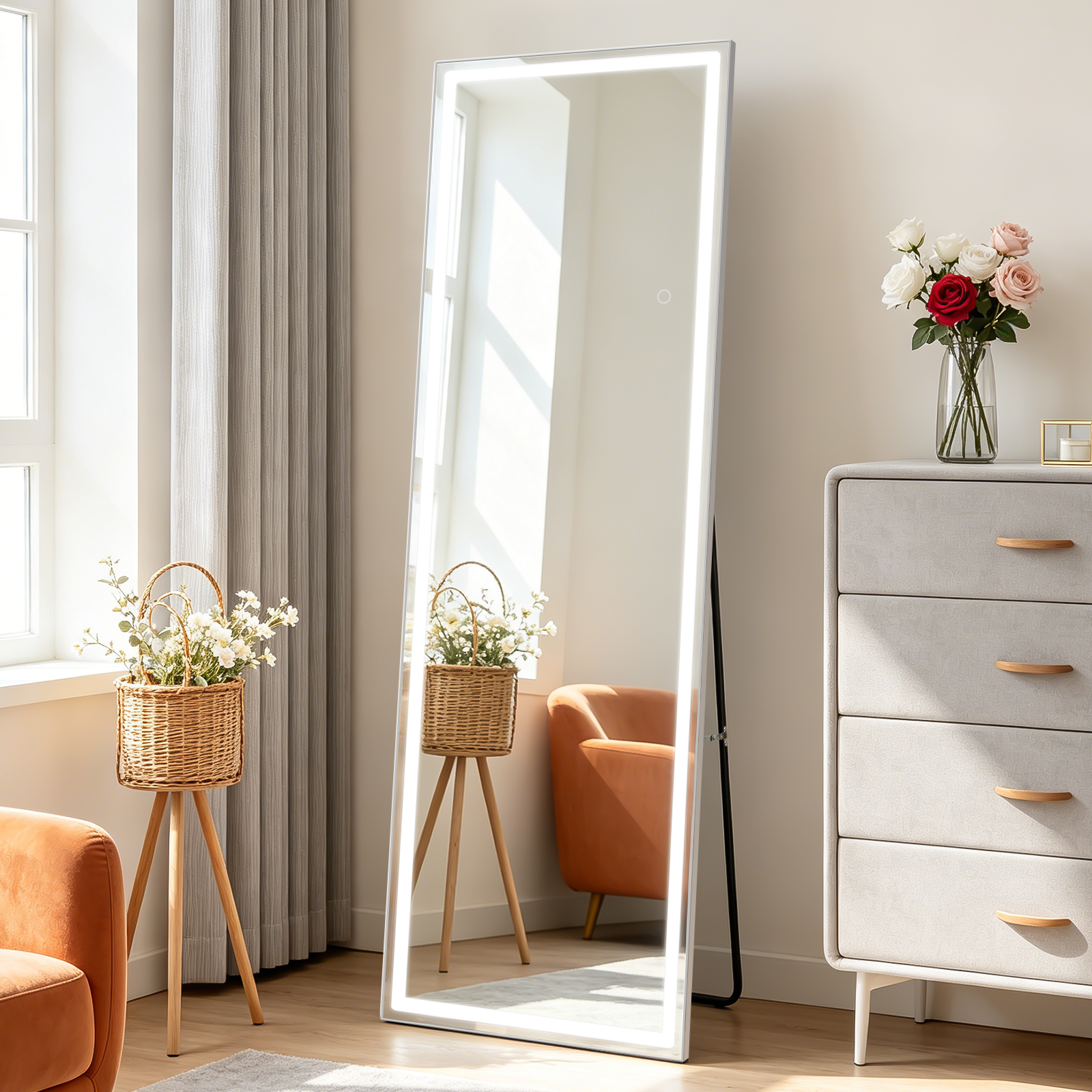 Un espejo LED de cuerpo entero se apoya en la pared junto a una cómoda gris, un sillón y una cesta con flores.