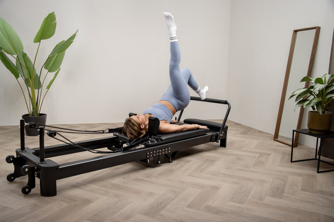 Una mujer rubia con ropa deportiva azul hace ejercicio en un reformador de pilates negro en una habitación luminosa.