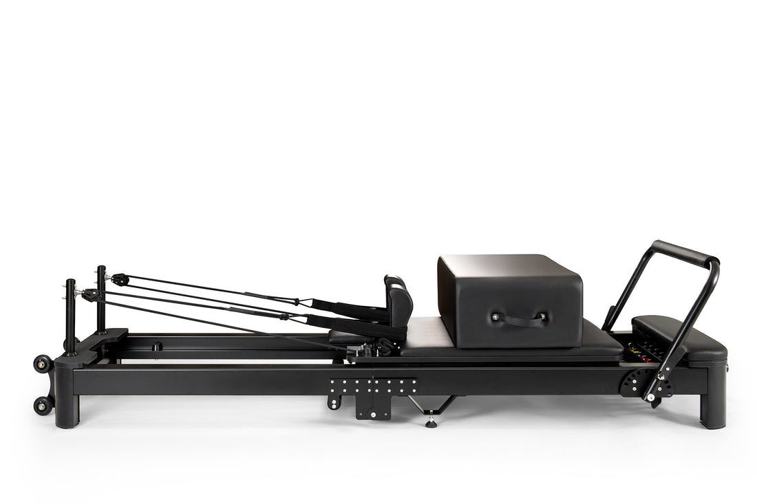 Una máquina reformer de pilates negra con carro acolchado, barra de pies, cuerdas y caja de salto sobre fondo blanco.