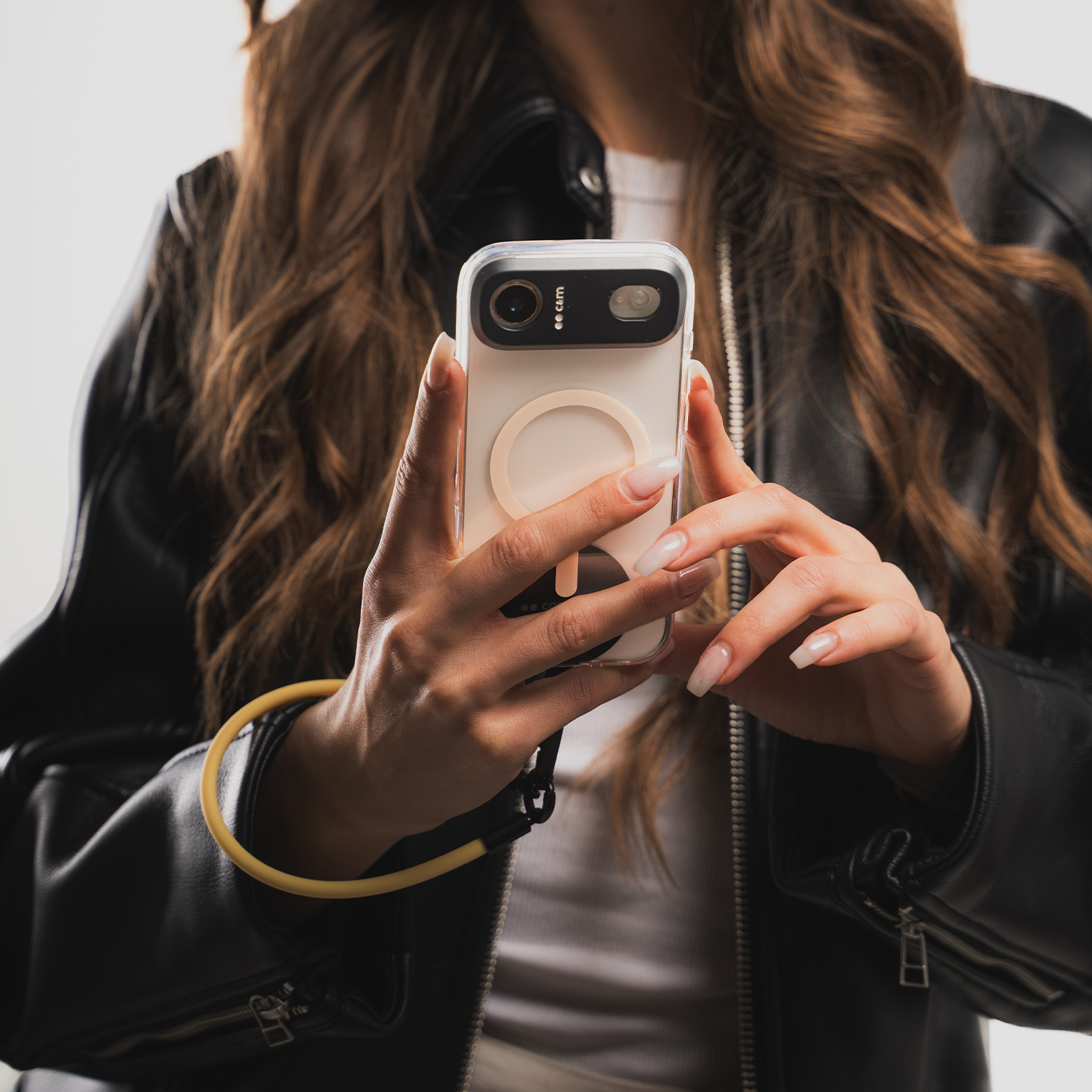 Una mujer con chaqueta de cuero sostiene un smartphone blanco con correa de muñeca amarilla.