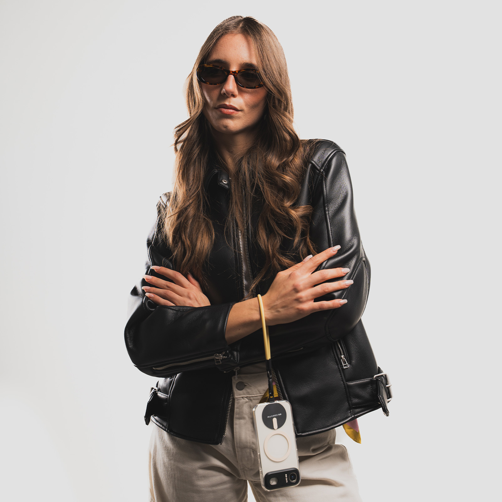Una mujer con cabello castaño largo, gafas de sol y chaqueta de cuero negra posa con los brazos cruzados ante pared blanca.