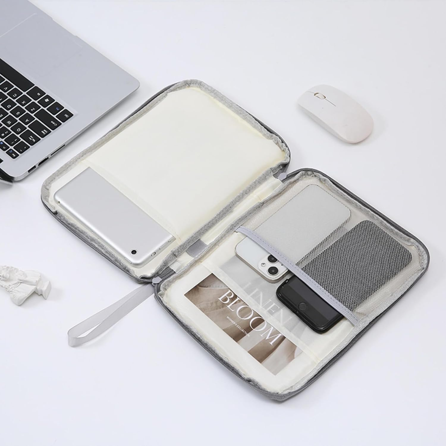 Offene graue Tasche mit elektronischen Geräten, ein Laptop und eine Maus liegen auf weißem Untergrund.