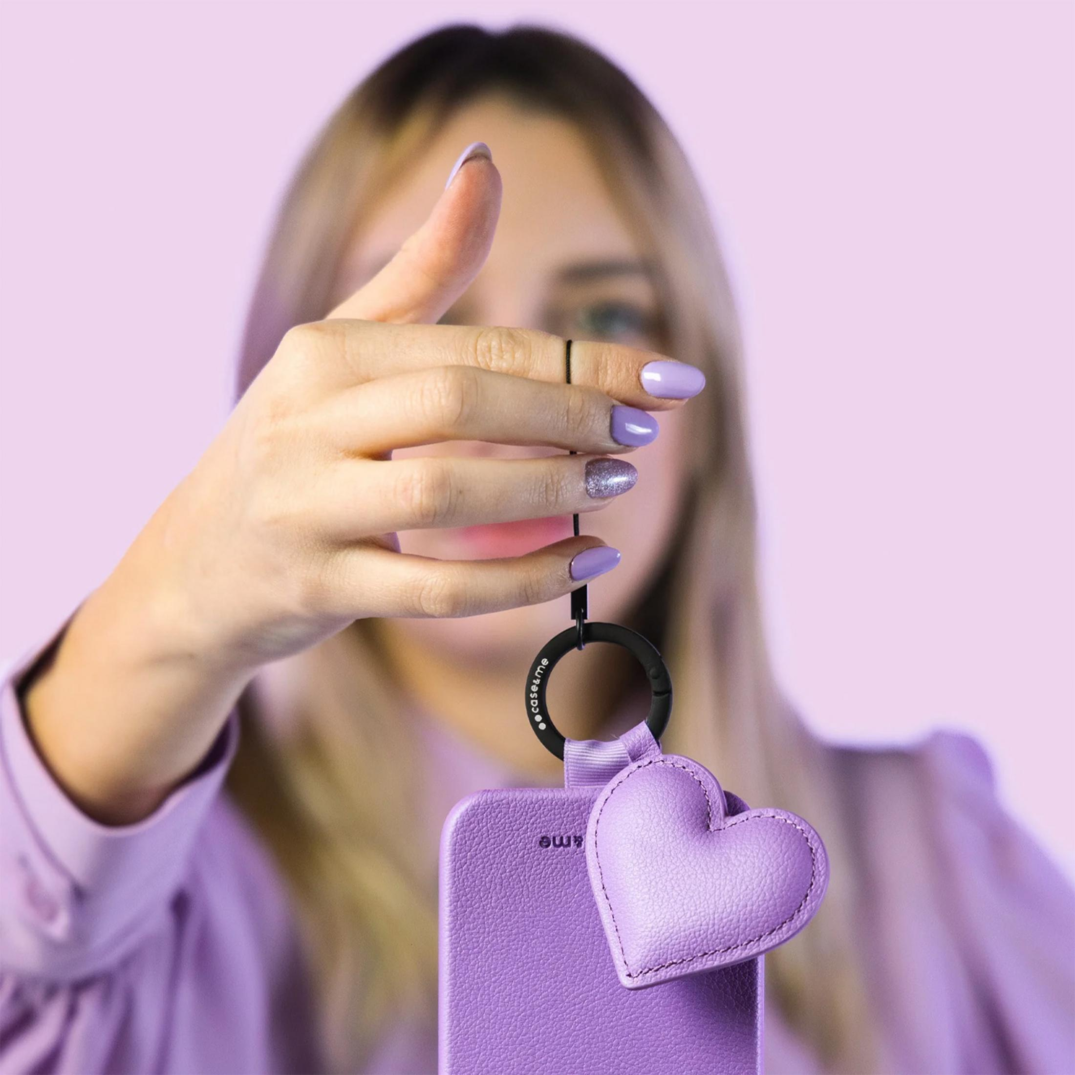 Una mujer rubia sostiene una funda morada con un colgante de corazón sobre fondo morado.