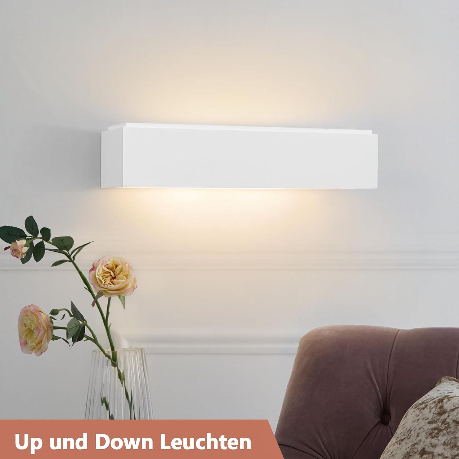 Un aplique blanco emite luz cálida hacia arriba y hacia abajo, junto a flores y un sillón.