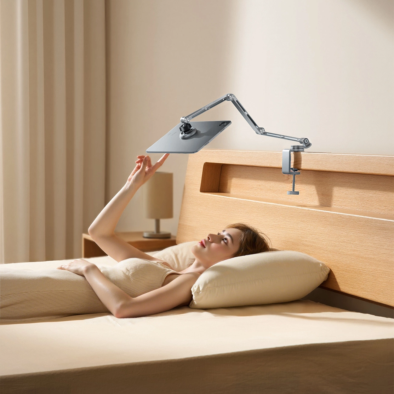 Una donna è sdraiata a letto e tocca un tablet tenuto da un braccio regolabile montato sulla testiera di legno.