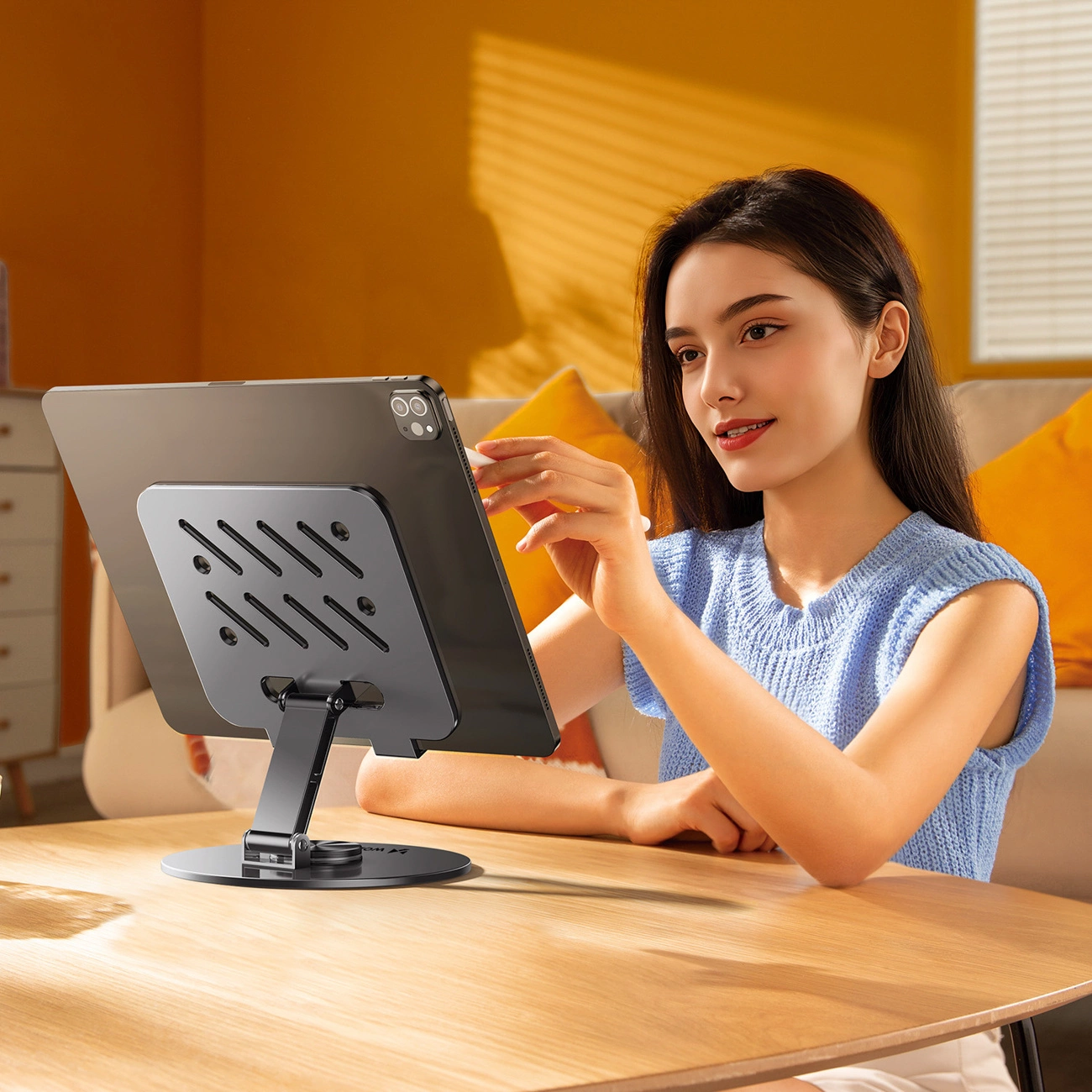 Una donna con un top blu siede a un tavolo di legno e interagisce con un tablet su un supporto.
