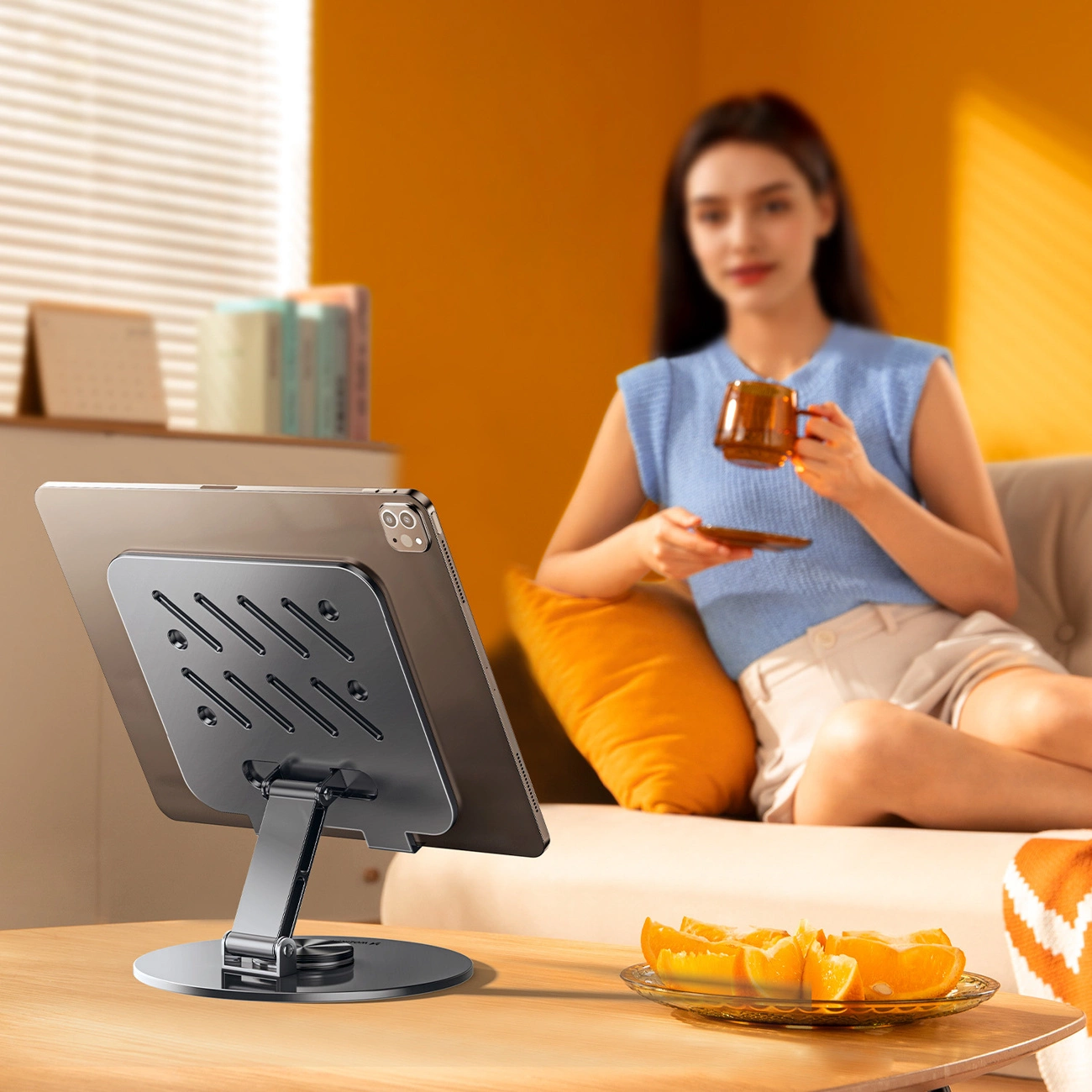 Un tablet su supporto con fette d'arancia sul tavolo e una donna con tazza sul divano sullo sfondo.