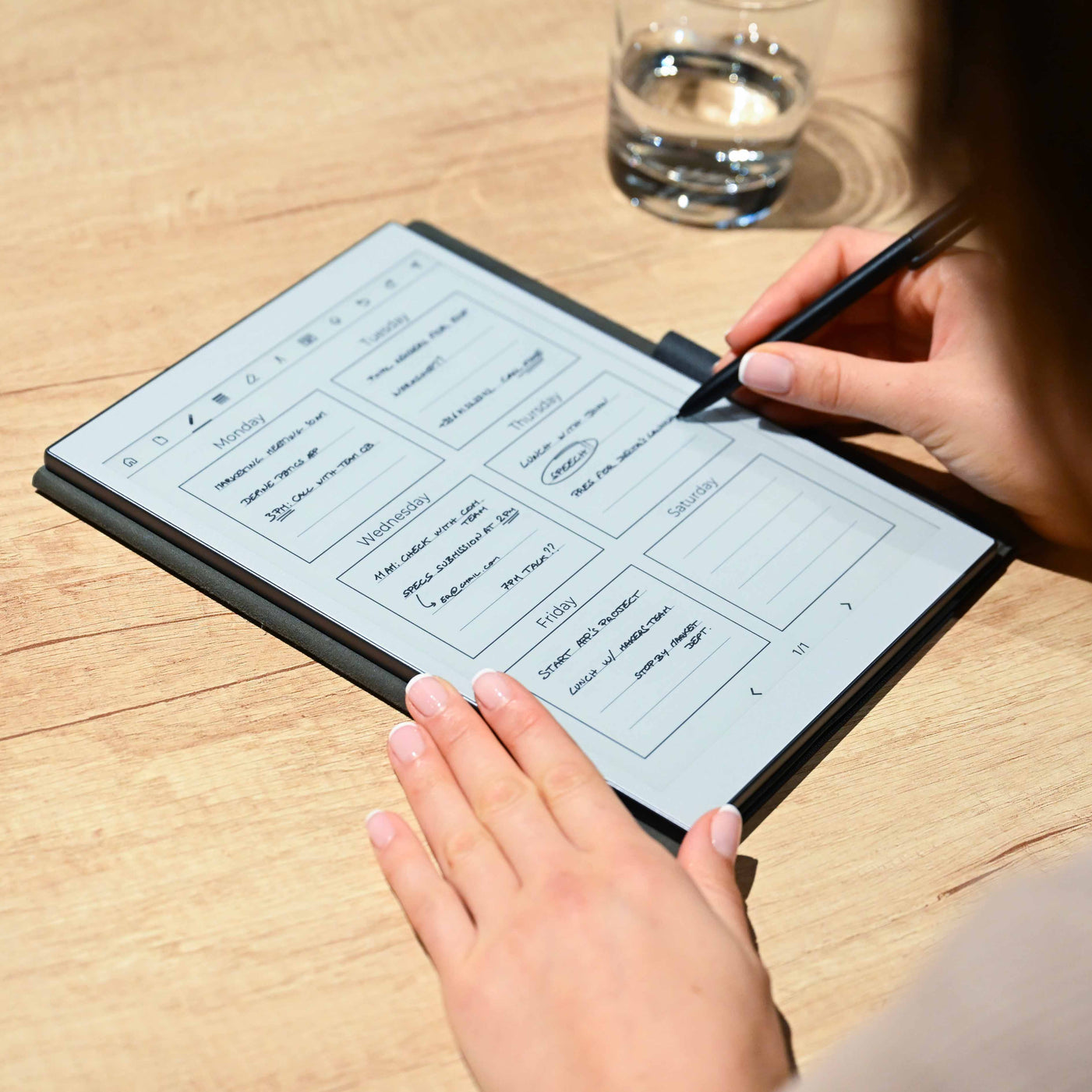 Persona escribe con bolígrafo negro en una tablet digital con calendario en mesa de madera.