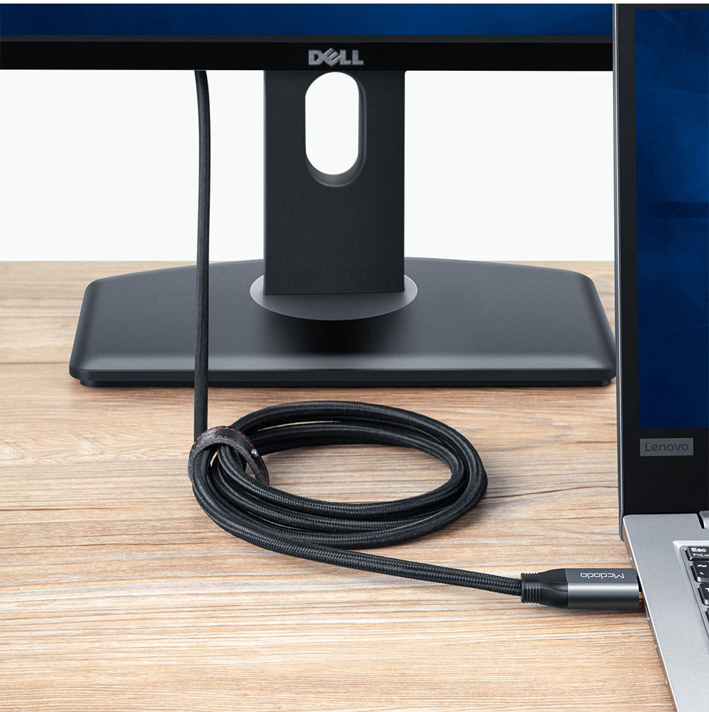 Un cavo nero intrecciato è arrotolato su una scrivania di legno tra un monitor e un computer portatile.