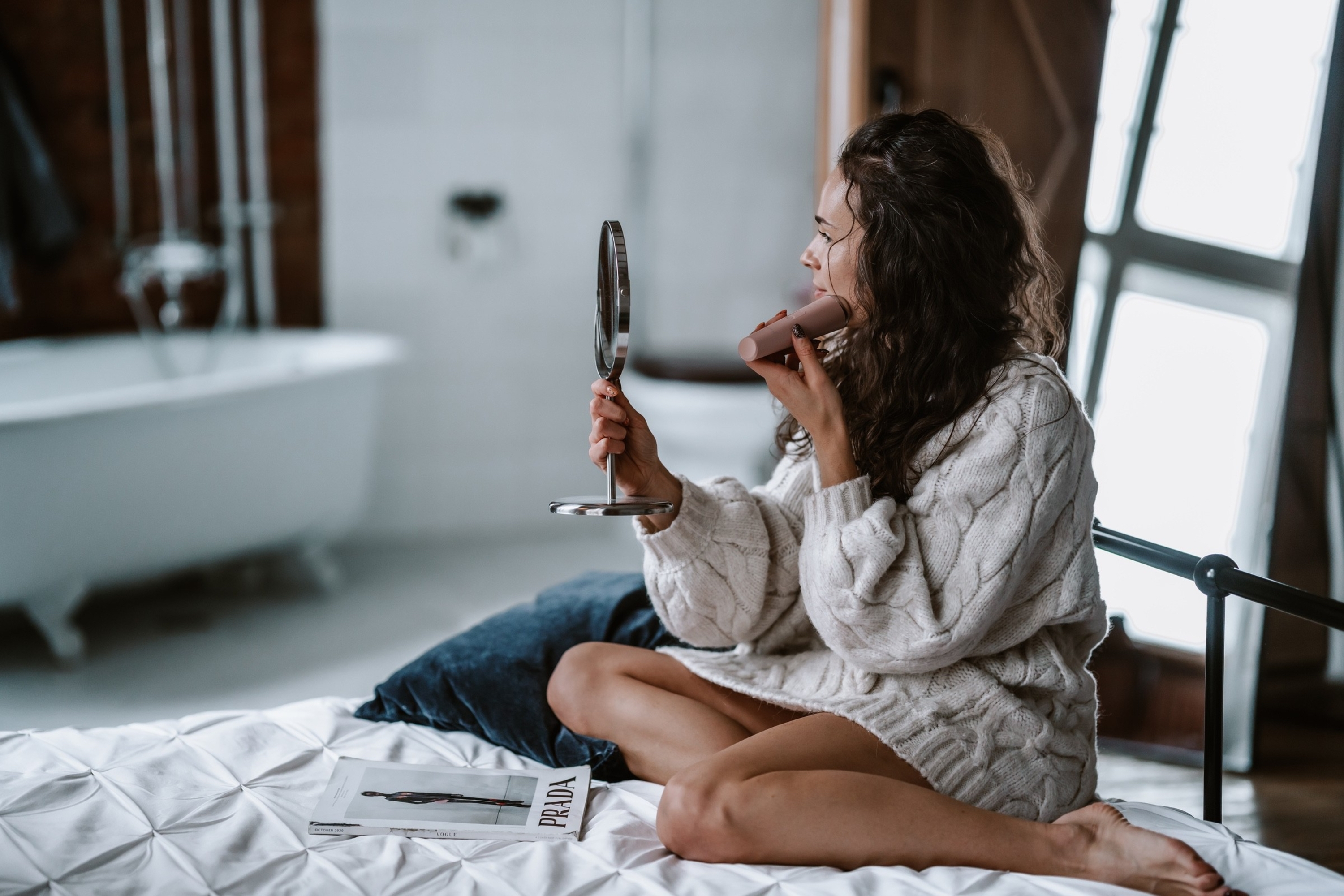 Donna seduta sul letto, tiene un piccolo specchio e si trucca le labbra.