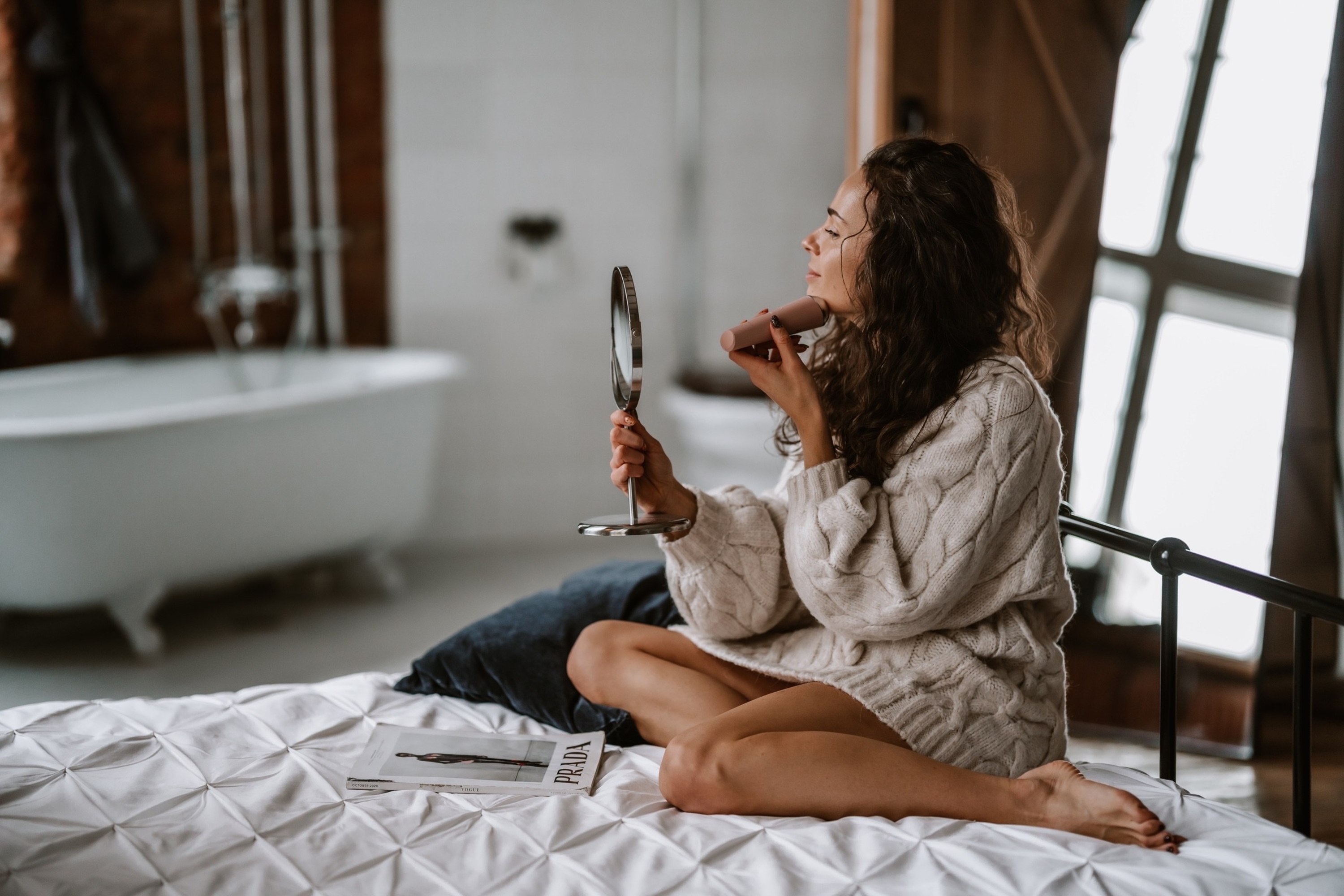 Una donna con maglione bianco siede sul letto, tiene uno specchio a mano e applica la crema.