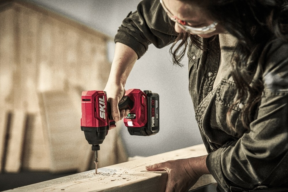 Una donna con occhiali protettivi usa un trapano a batteria rosso per avvitare una tavola di legno.