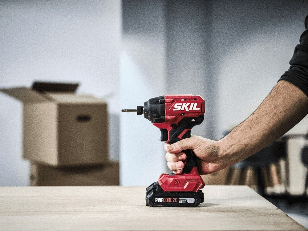 Una persona tiene in mano un trapano a batteria Skil rosso e nero su un tavolo di legno, con scatoloni sullo sfondo.