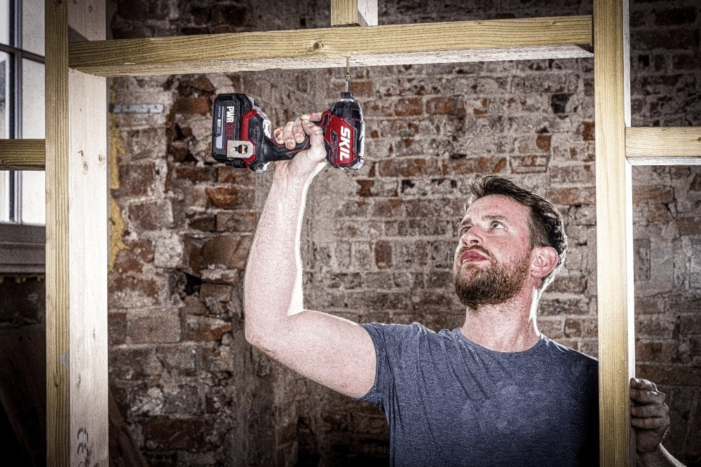 Un uomo con la barba usa un trapano a batteria su una struttura in legno contro un muro di mattoni.