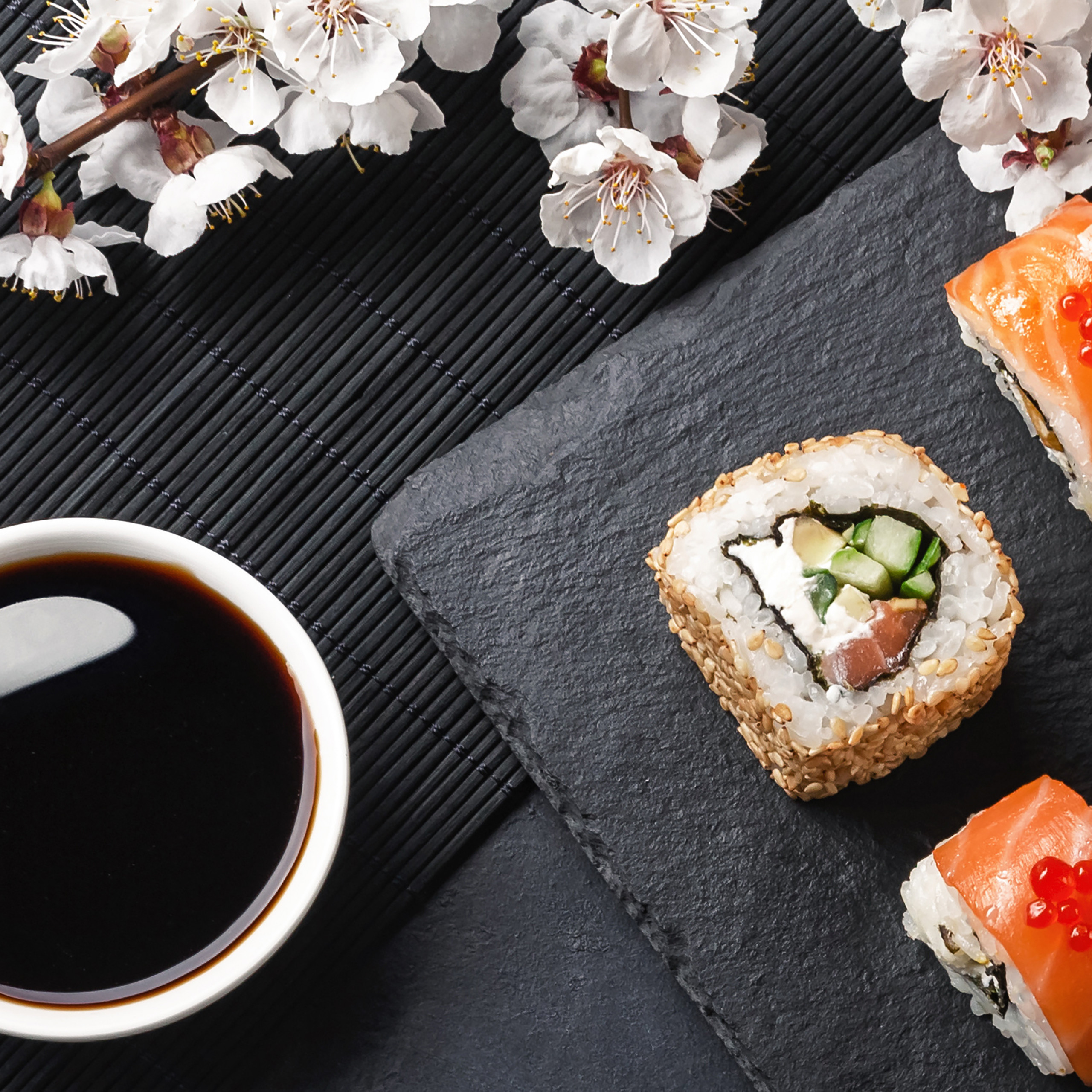 Sushi-Rollen auf einer Schieferplatte, eine Schale Sojasauce und weiße Blüten auf einer schwarzen Bambusmatte.