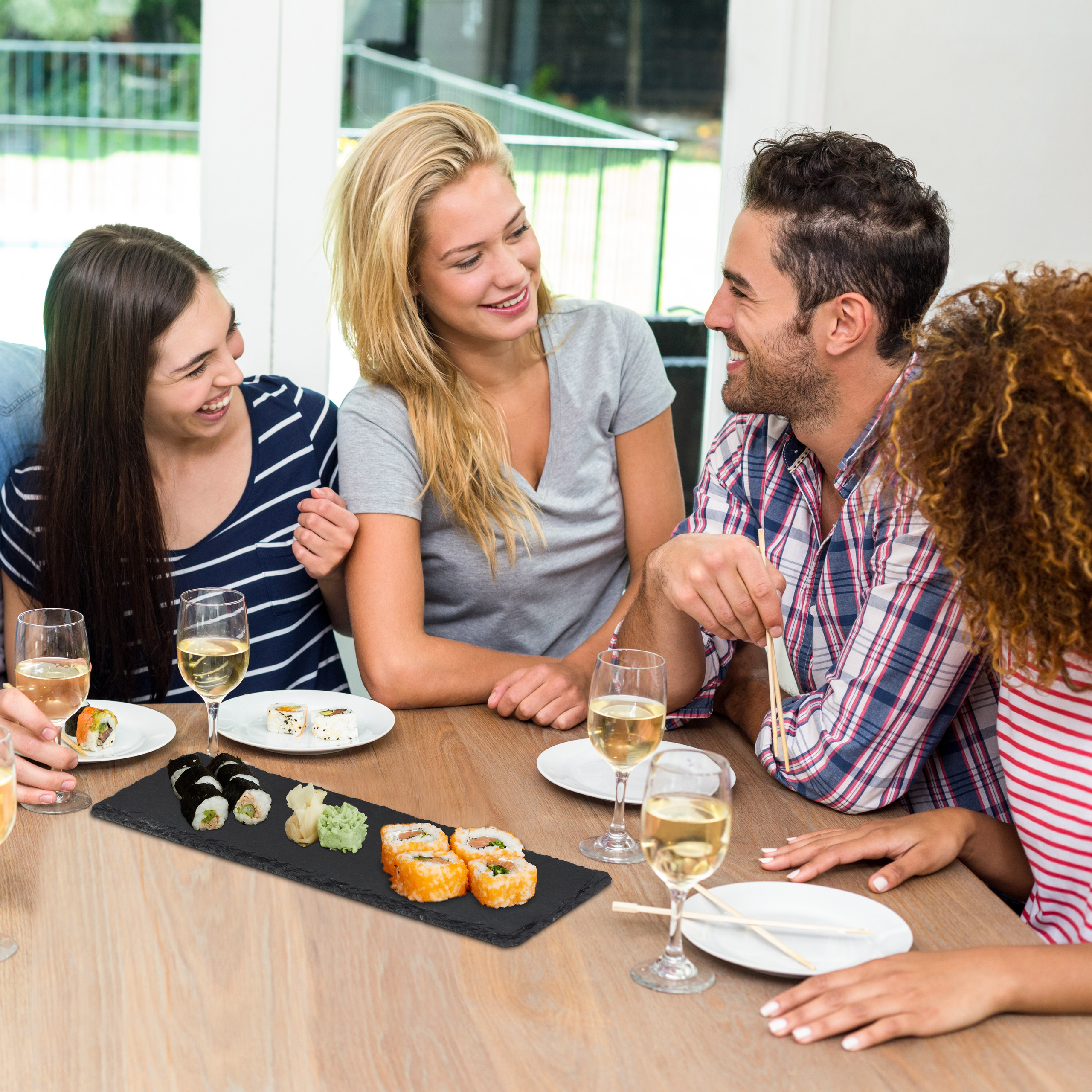 Vier Freunde lachen und essen Sushi an einem Holztisch mit Weingläsern.