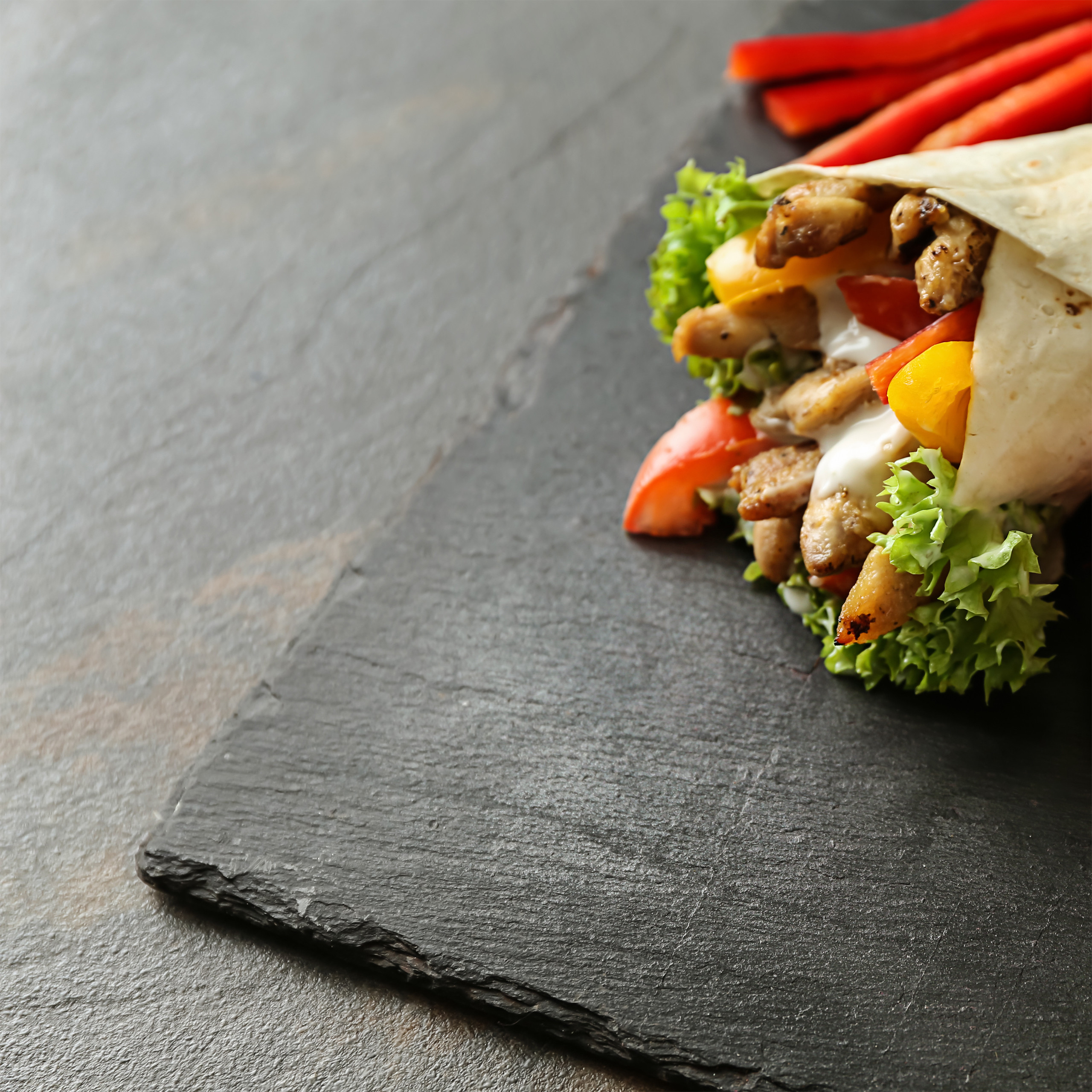 Ein Hähnchen-Wrap mit Salat, Paprika und Soße liegt auf einem dunklen Schieferbrett.