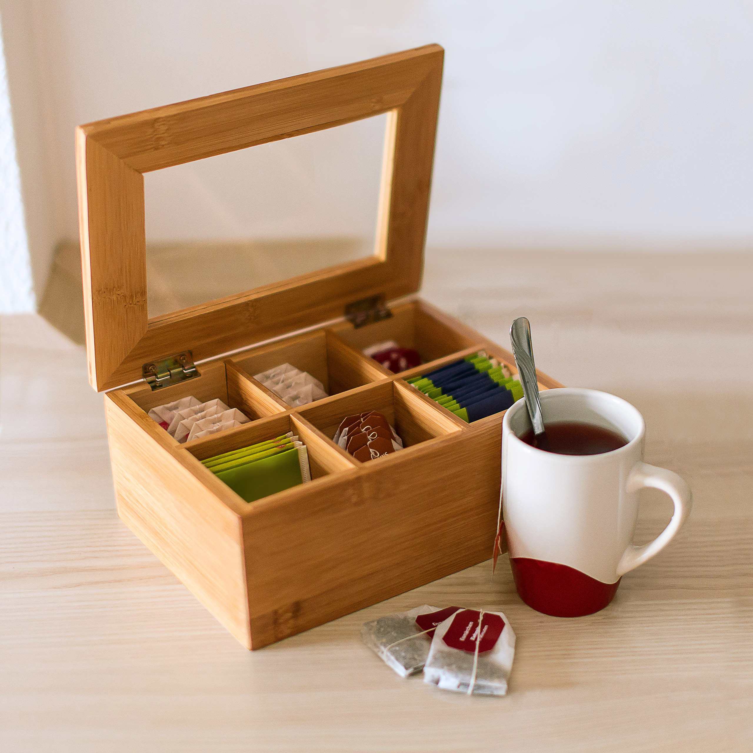 Eine offene Teebox aus Holz mit Teebeuteln steht neben einer weiß-roten Tasse mit Löffel auf einem Holztisch.