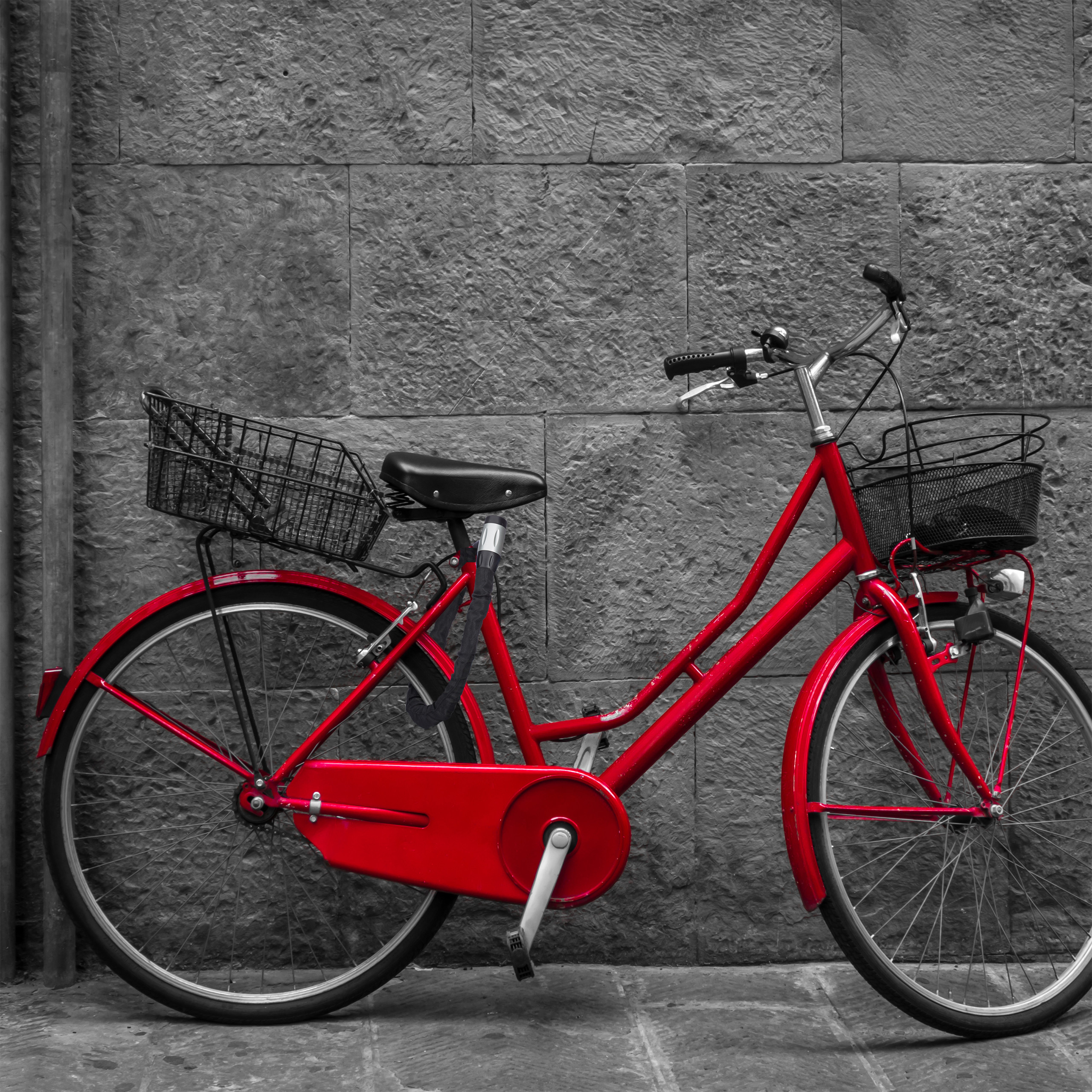 Ein leuchtend rotes Fahrrad mit Körben vorne und hinten lehnt an einer grauen Steinmauer in Schwarz-Weiß.