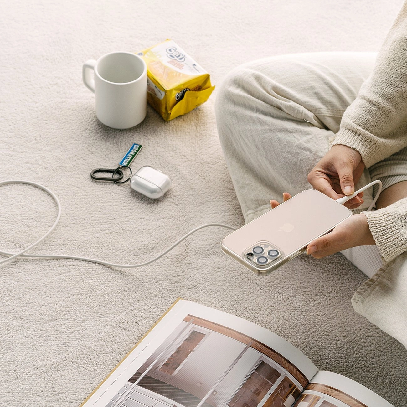 Una persona seduta su un tappeto collega un cavo a un telefono vicino a una tazza, snack e un libro aperto.