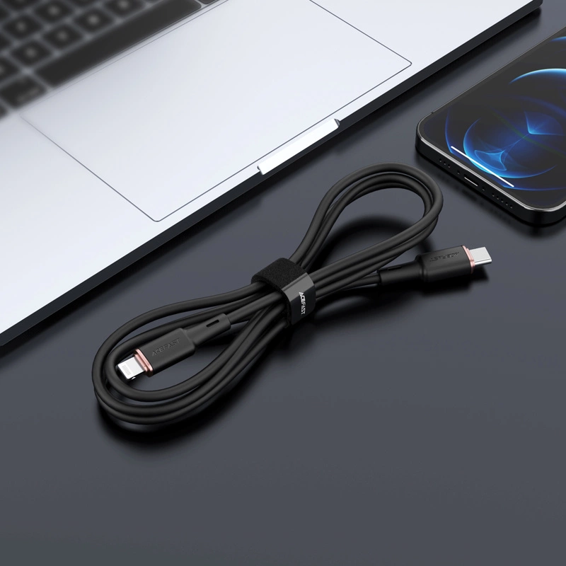 Un cavo di ricarica nero arrotolato si trova su una scrivania scura accanto a un laptop e uno smartphone.