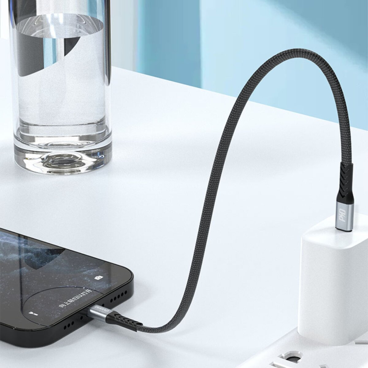 Un cavo di ricarica nero intrecciato collega uno smartphone a un caricabatterie da muro accanto a un bicchiere d'acqua.