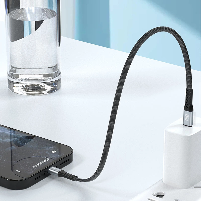 Un cavo di ricarica intrecciato nero collega uno smartphone a un caricabatterie da muro accanto a un bicchiere d'acqua.