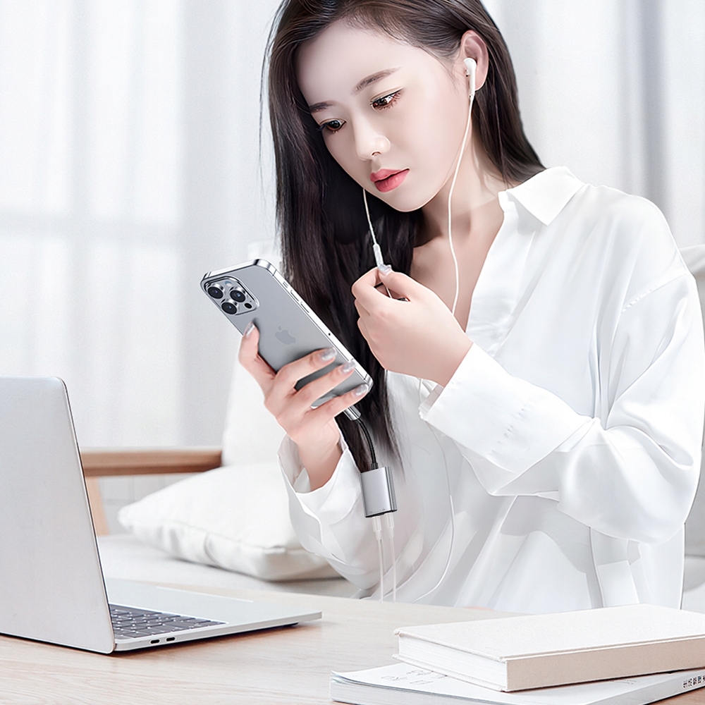 Una donna in camicia bianca siede a un tavolo con un laptop, tiene uno smartphone con adattatore e indossa auricolari.