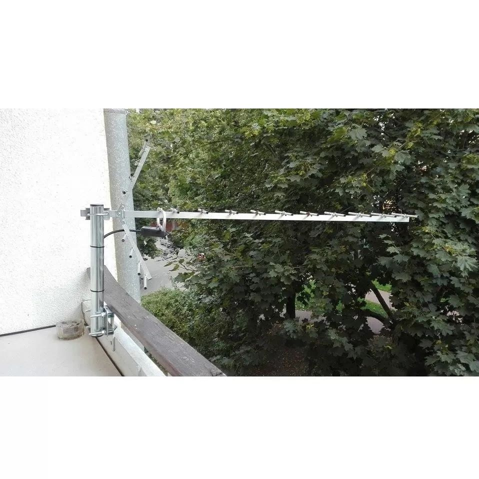 Eine lange Metallantenne ist an einem Balkongeländer vor grünen Bäumen montiert.