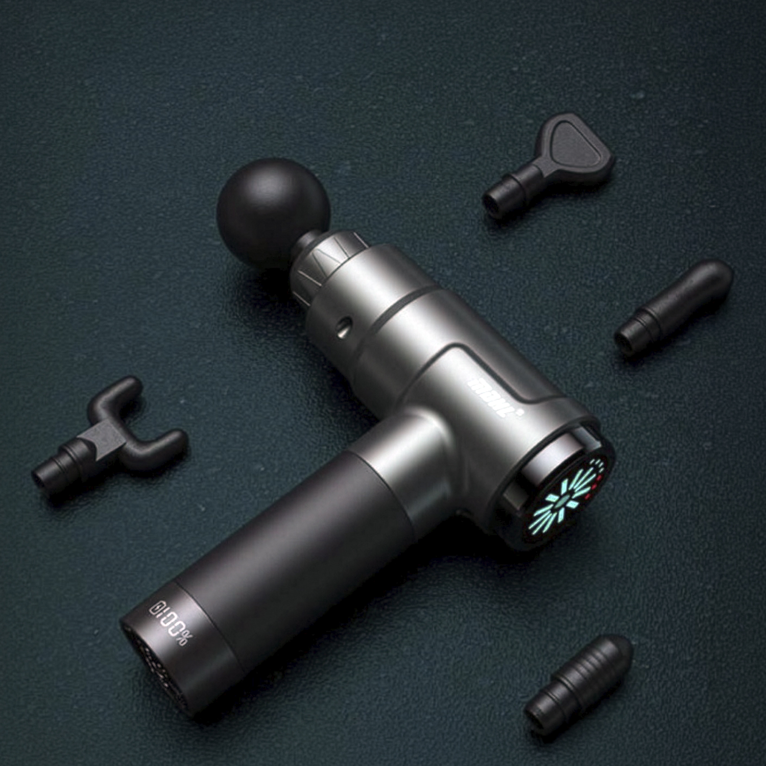 Una pistola de masaje gris yace sobre una superficie oscura, rodeada de cuatro accesorios.