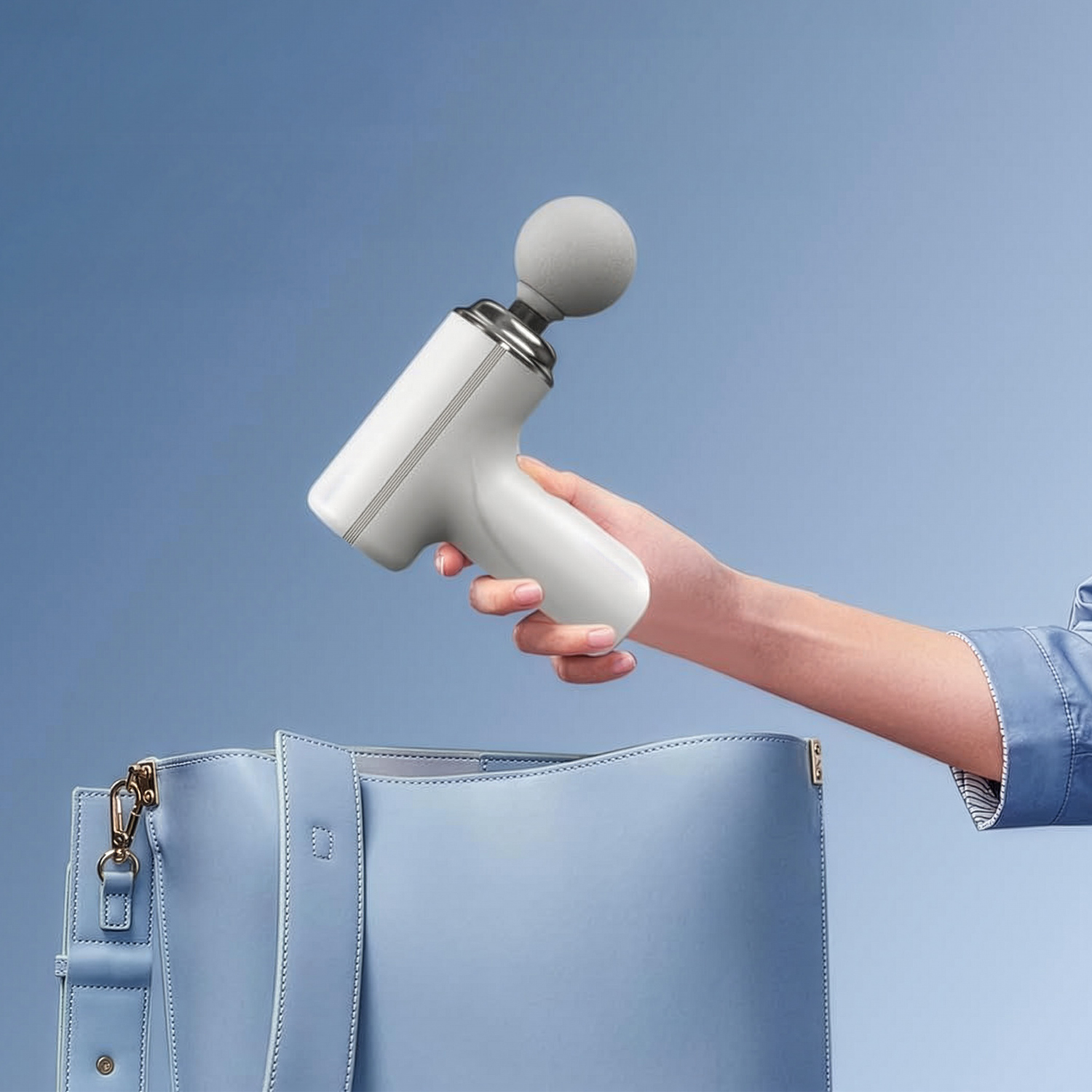 Una mano sostiene una pequeña pistola de masaje gris y blanca sobre un bolso azul claro con fondo azul.