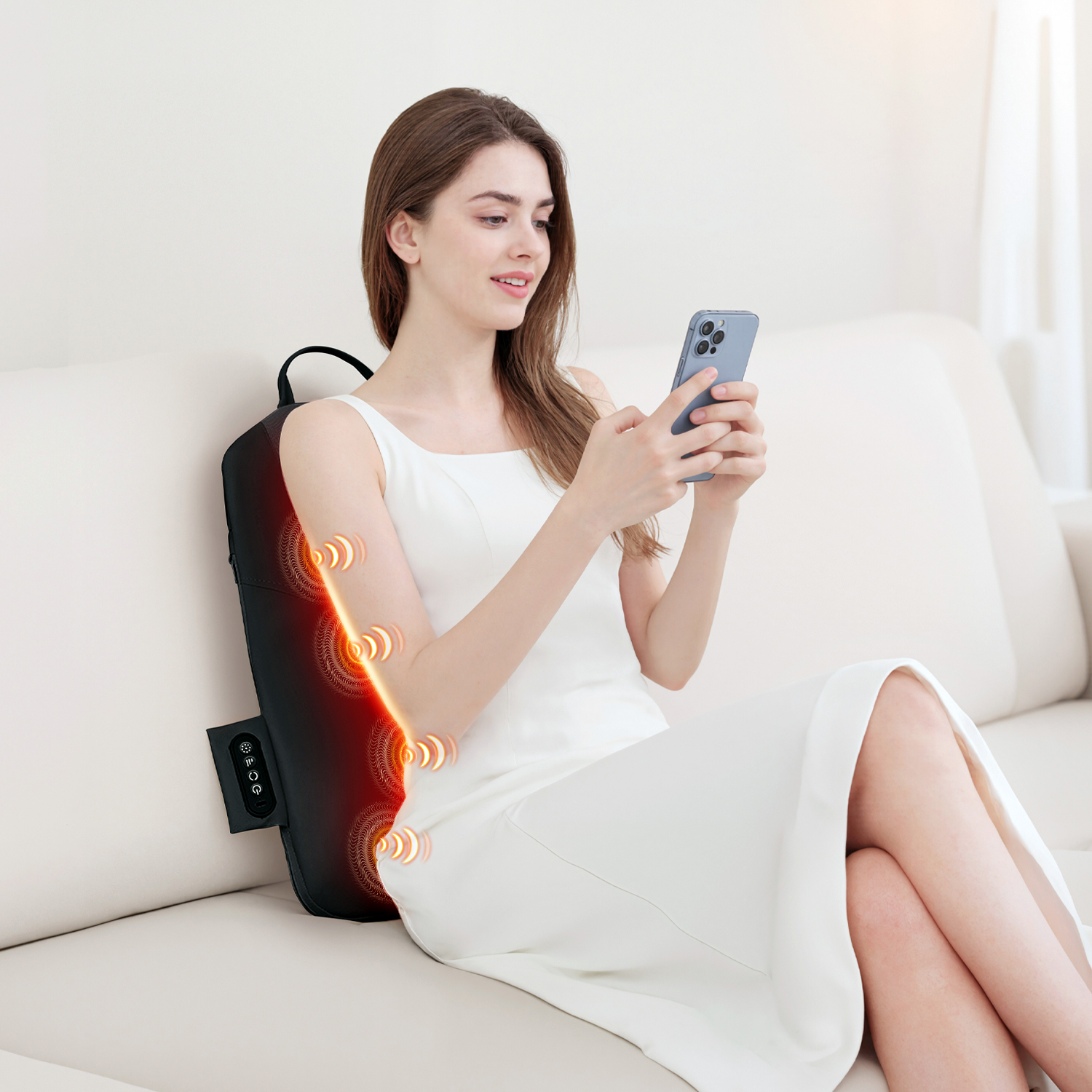 Una mujer con un vestido blanco se sienta en un sofá, usando su teléfono inteligente mientras un masajeador de espalda negro se coloca detrás de ella.