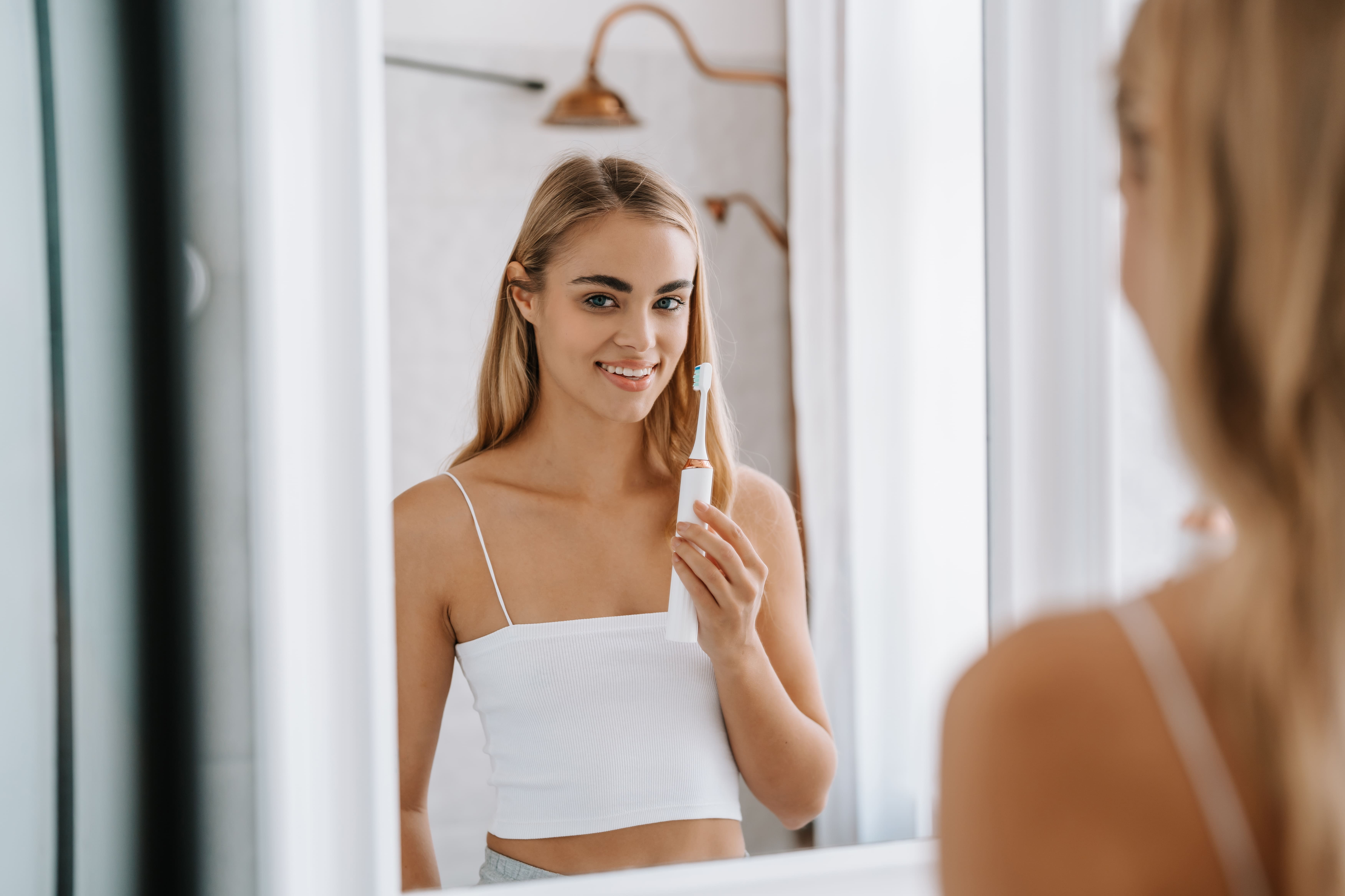 Blond kobieta uśmiecha się przed lustrem, trzymając elektryczną szczoteczkę do zębów w jasnej łazience.