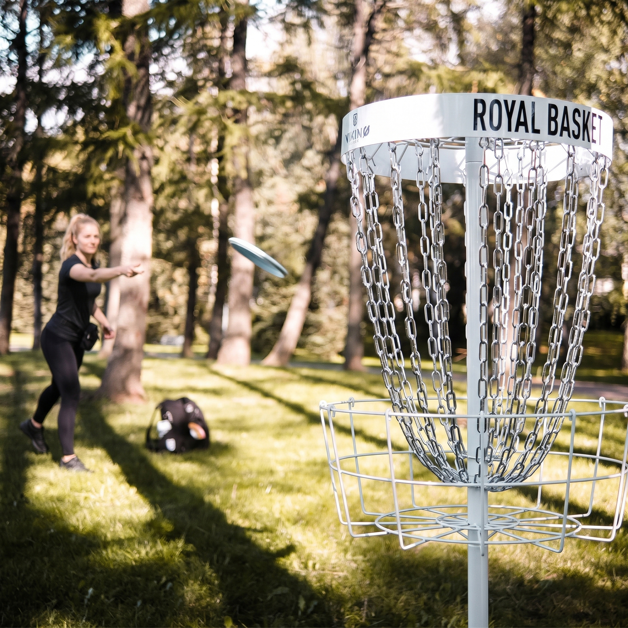 Eine Frau wirft eine Frisbee-Scheibe in Richtung eines königlichen Disc-Golf-Korbs in einem bewaldeten Park.