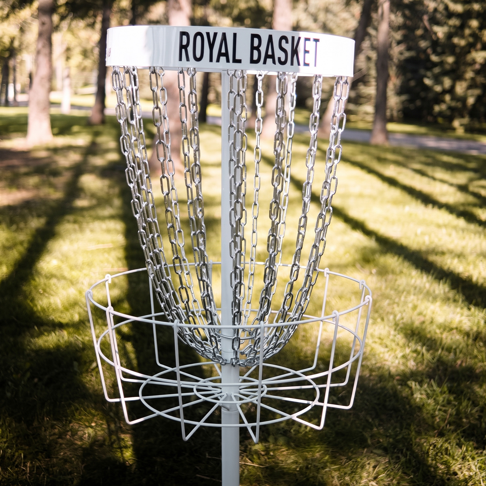 Ein weißer Disc-Golf-Korb mit Metallketten steht auf einer sonnigen Wiese vor Bäumen.