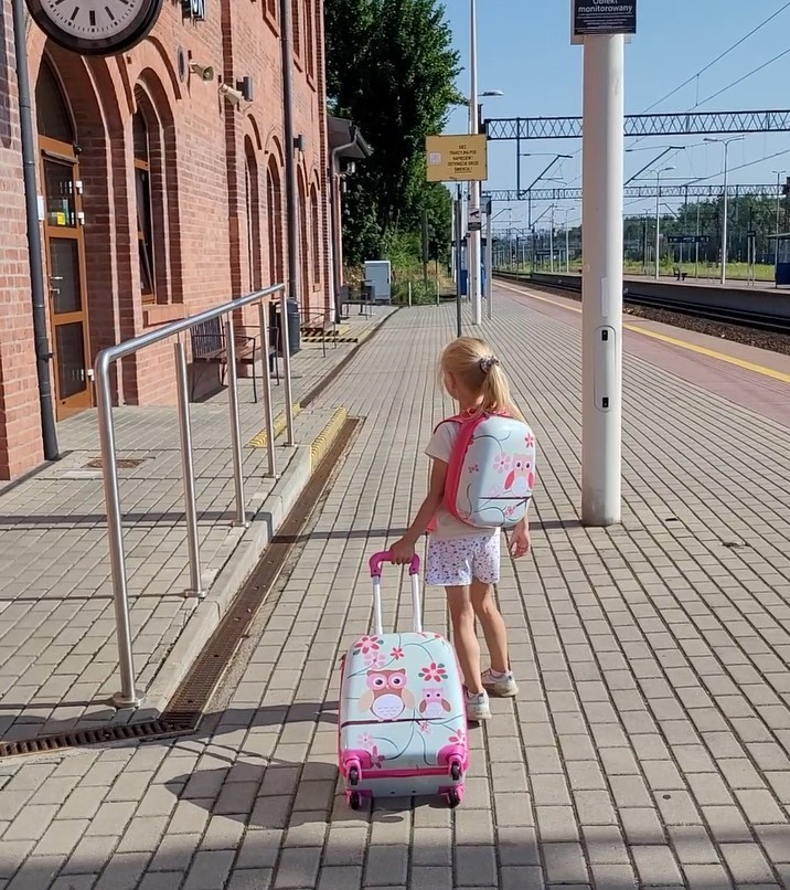 Dziewczynka z pasującym plecakiem w sowy i walizką na kółkach idzie po peronie ceglanej stacji kolejowej.