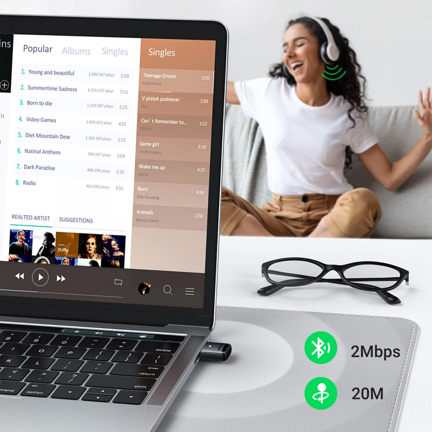 Laptop con interfaccia musicale, adattatore wireless, occhiali neri sul tavolo e donna che balla sullo sfondo.