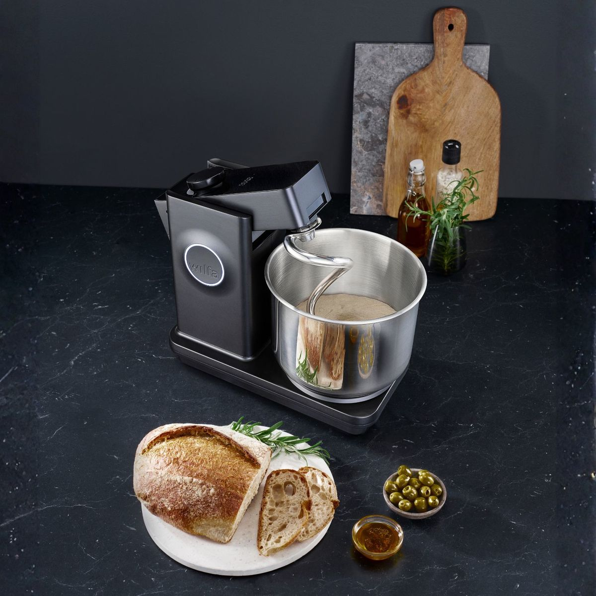 Una impastatrice nera su un bancone con pane, olio d'oliva, olive e un tagliere di legno sullo sfondo.