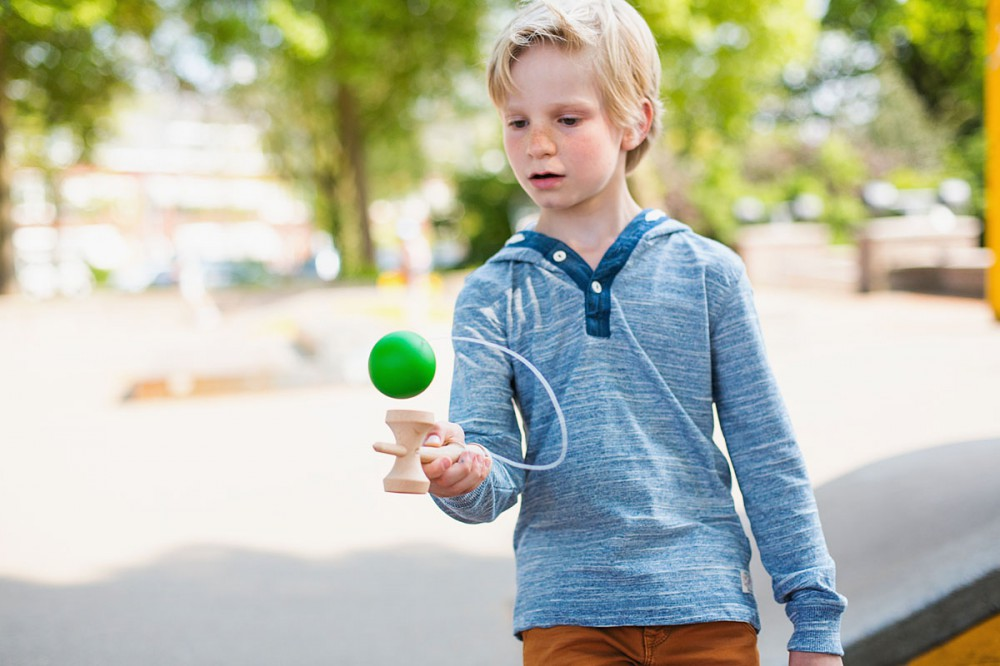 Ein junger blonder Junge spielt draußen in einem Park mit einem Kendama-Holzspielzeug mit grünem Ball.