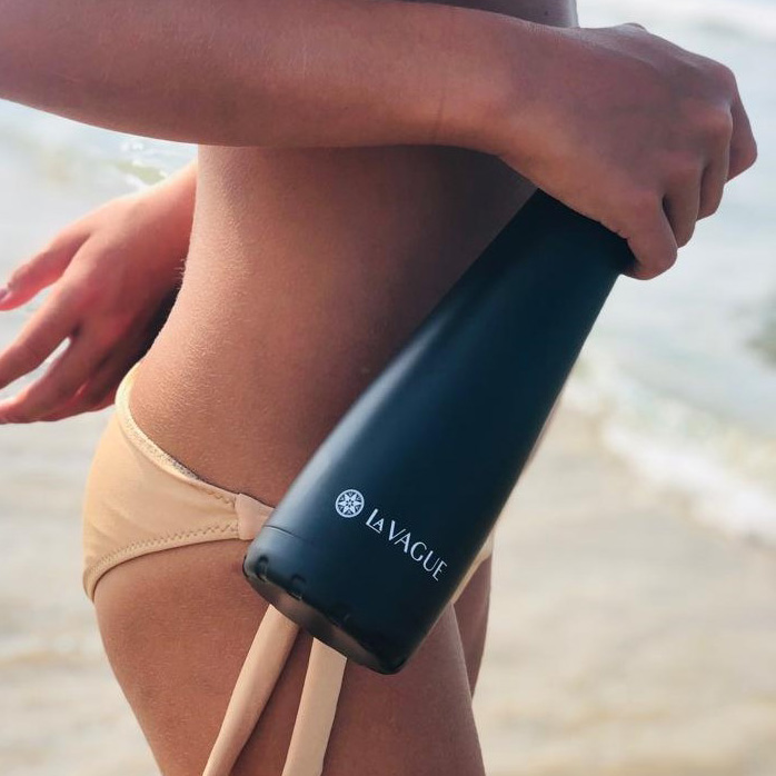 Eine Frau in beigem Bikini hält eine schwarze "La Vague" Trinkflasche am Strand.