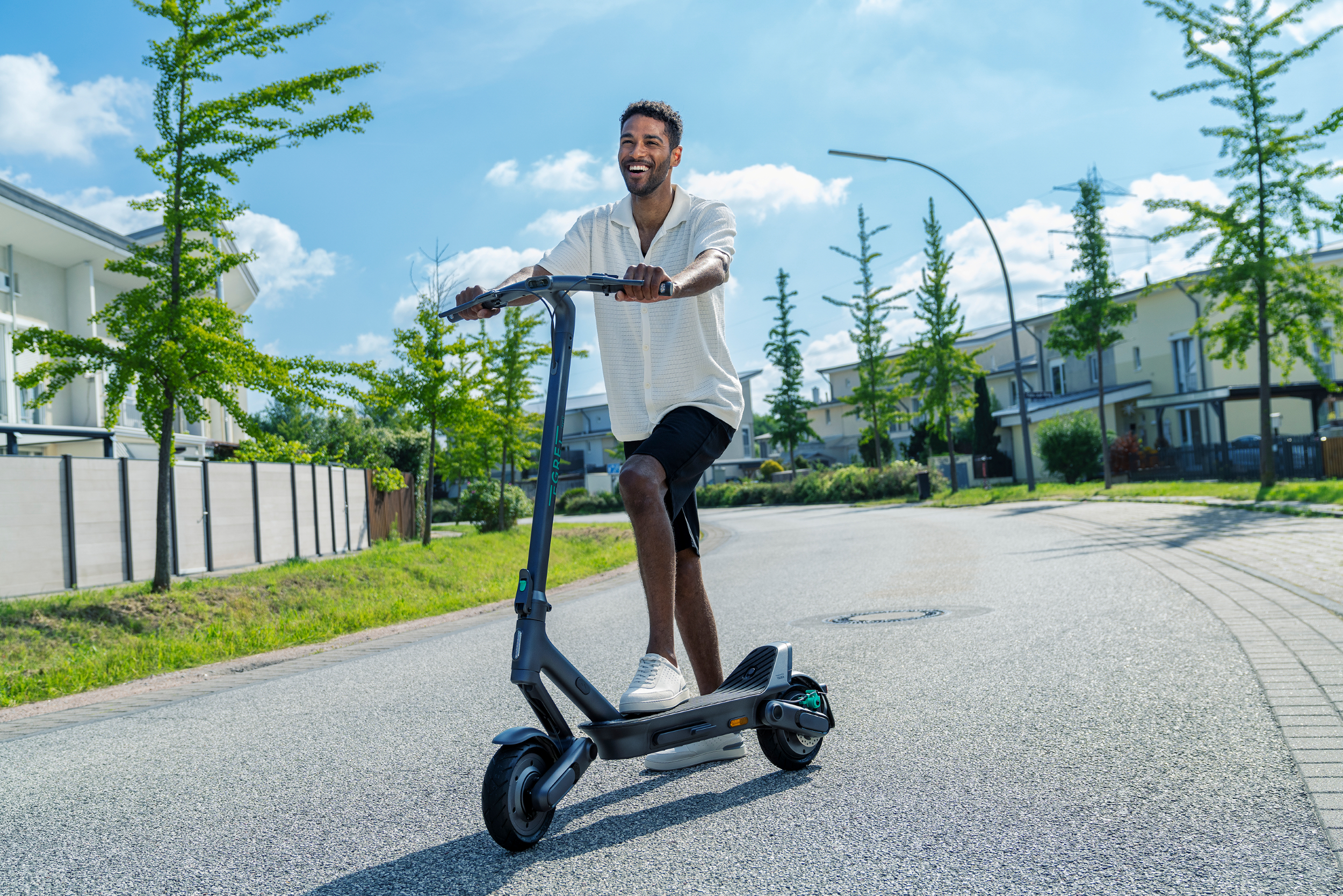 Ein lächelnder Mann in weißem Hemd und schwarzen Shorts fährt an einem sonnigen Tag mit einem E-Scooter durch eine Straße.