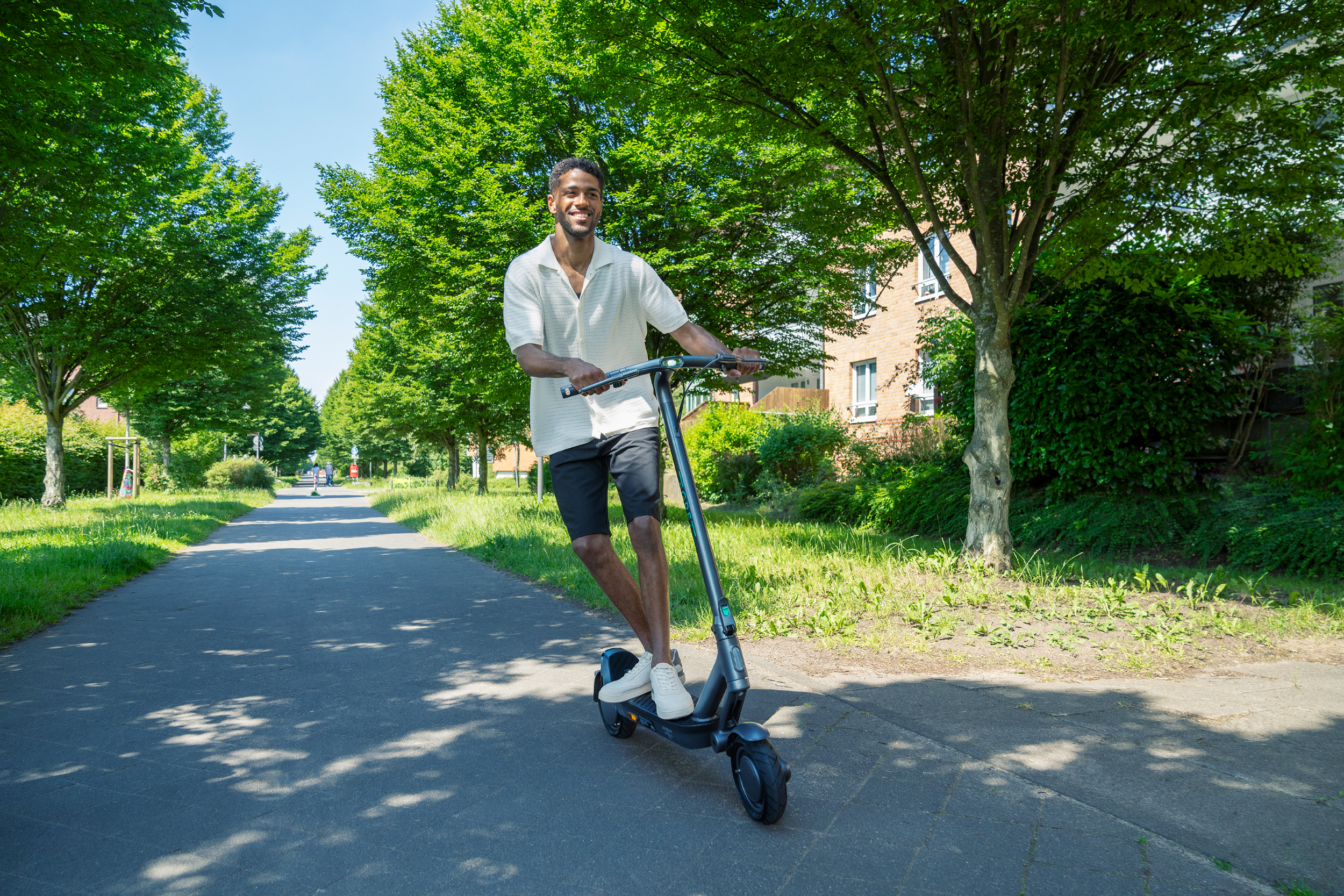 Ein junger Mann lächelt, während er an einem sonnigen Tag auf einem E-Scooter auf einem von Bäumen gesäumten Weg fährt.