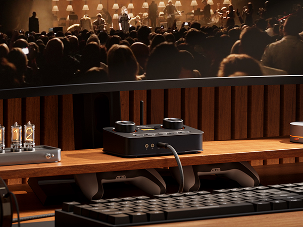 Un mixer audio con manopole su una scrivania in legno, davanti a uno schermo che mostra una folla.