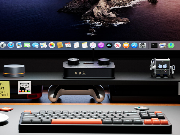 Una scrivania con monitor, tastiera, dispositivo audio, gamepad e statuetta di robot.
