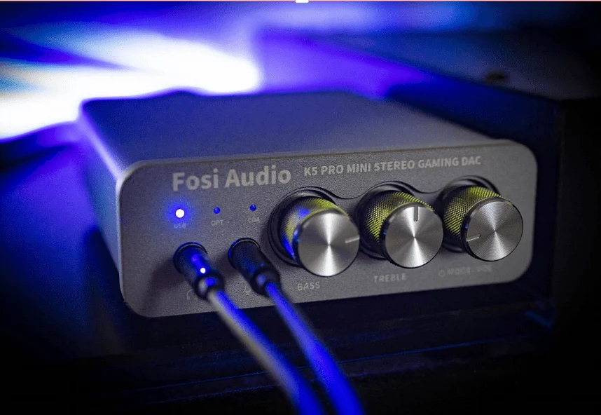 Fosi Audio K5 Pro gaming DAC with three knobs, blue LED light, and two connected cables against a blue background.