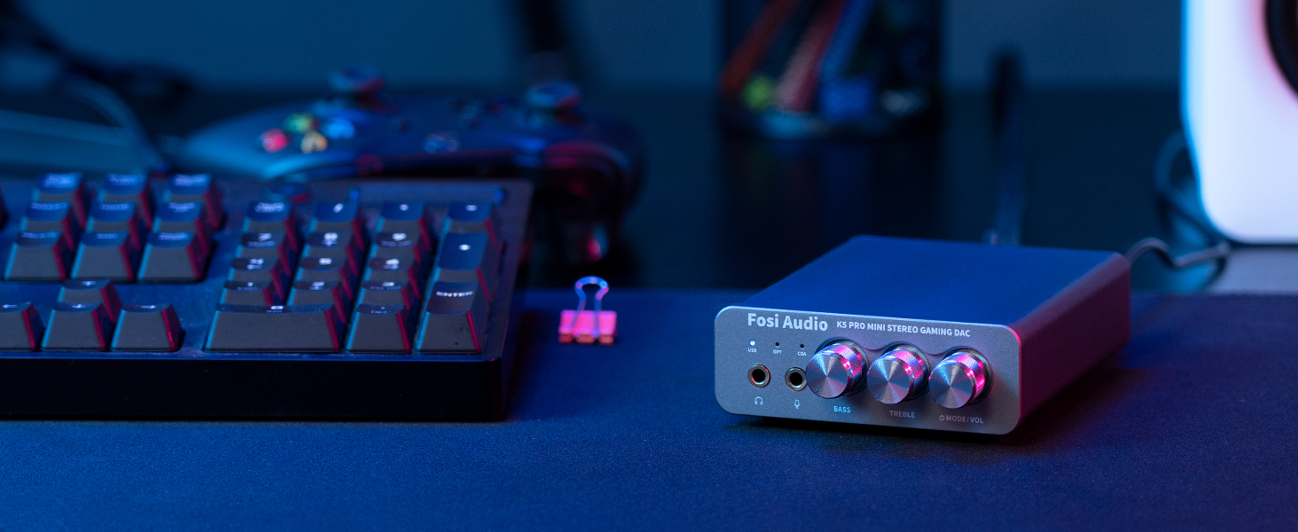 Przetwornik DAC Fosi Audio K5 Pro stoi na niebiesko oświetlonym biurku obok klawiatury i kontrolera.
