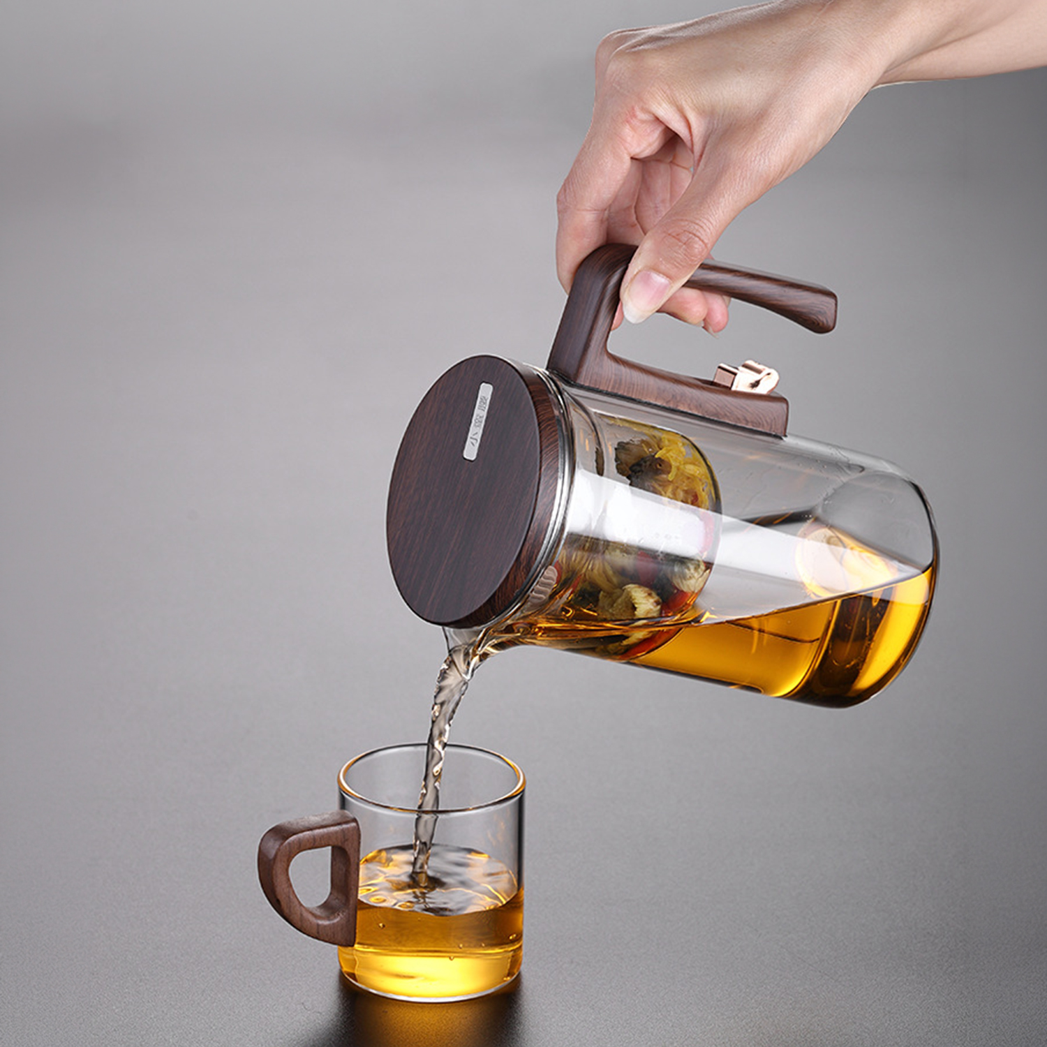 Una mano vierte té de una tetera de cristal con asa y tapa de madera en una taza pequeña de cristal.