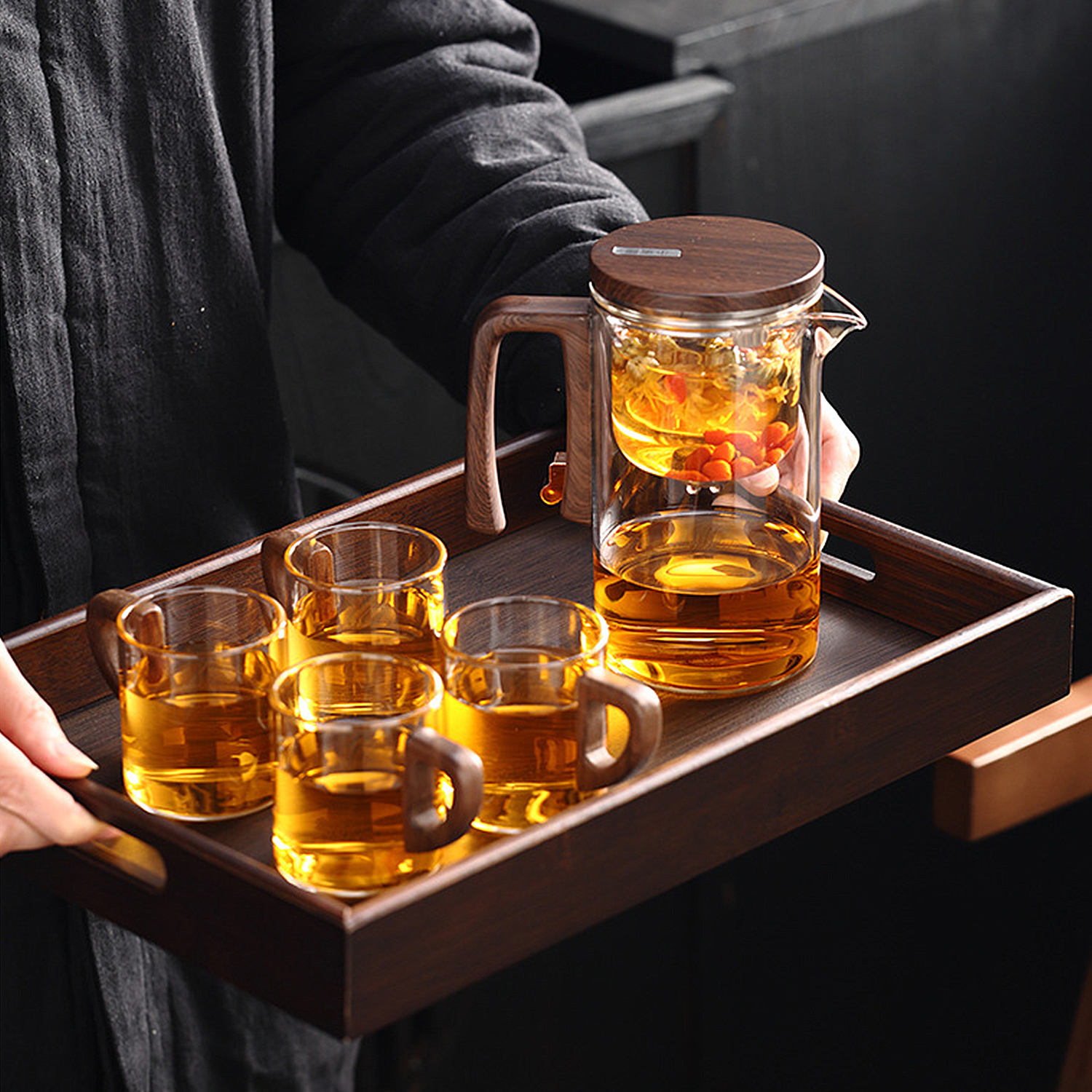 Una persona sostiene una bandeja de madera oscura con una tetera de vidrio y cuatro tazas pequeñas con té ámbar.